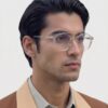 A man with glasses wearing a brown jacket, standing against a neutral background.