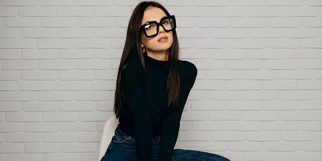 A woman with glasses sits comfortably on a chair, looking thoughtfully to the side.