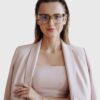 A woman wearing glasses and a pink suit, smiling confidently while standing against a neutral background.
