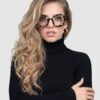 A beautiful blonde woman in glasses wearing a black turtleneck, smiling softly at the camera.