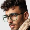 A man wearing glasses and a stylish fur coat, posing confidently against a neutral background.