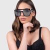 A woman wearing black sunglasses and a black bra top, posing confidently against a neutral background.