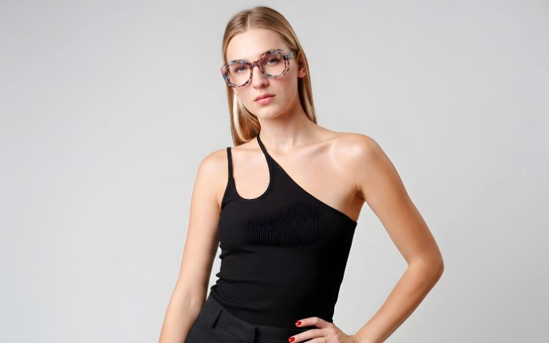 A woman wearing black glasses and a one-shoulder top, posing confidently against a neutral background.